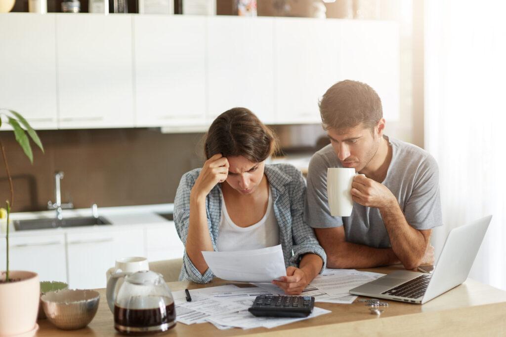 Female and male sitting together against kitchen interior, having attentive look into documents, being busy with managing finances, drinking coffee. Young successful business people coworking at home